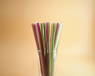 A collection of colorful straws stands upright in a clear glass against a warm, beige background. The straws vary in hues, including purple, green, and beige, and are arranged closely together.