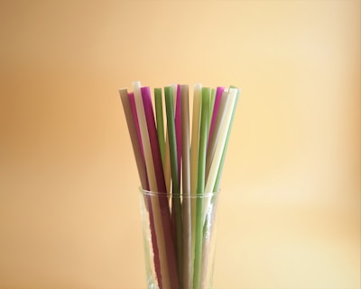 A collection of colorful straws stands upright in a clear glass against a warm, beige background. The straws vary in hues, including purple, green, and beige, and are arranged closely together.