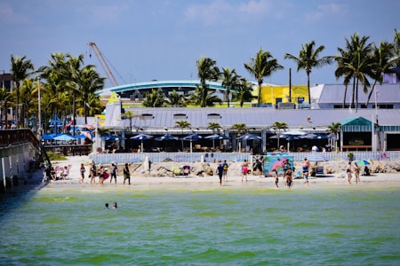 A lively beach scene with people walking and relaxing along the sandy shore. Palm trees line the background, framing a beachside building with colorful accents. Bright umbrellas and beachwear add splashes of color to the scene. In the foreground, two individuals are wading in the greenish-blue water.