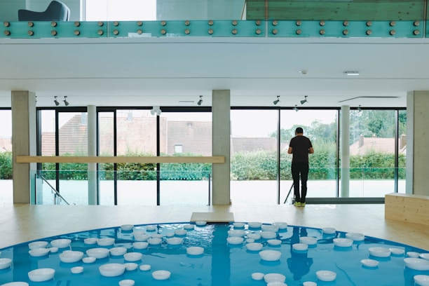 A modern indoor space with large glass windows looking out onto a garden. The foreground features a circular pool of blue water with floating white bowls. A person stands near the windows, looking outside. The upper area includes a glass railing with wooden accents, and a modern chair is visible in the top left corner.
