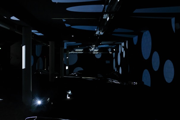 A dark room filled with large circular patterns projected onto the walls and ceiling. The room has several vertical pillars and scattered equipment. Soft blue and black hues dominate the space, creating an abstract and mysterious atmosphere.