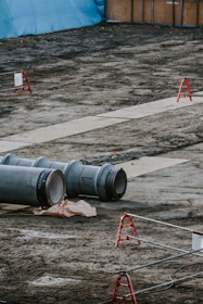 A construction site featuring large metal pipes resting on the ground, with red barriers scattered across the area. In the background, a blue tarp and wooden panels form a temporary wall. The ground is bare and appears recently disturbed.