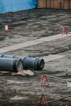 A construction site featuring large metal pipes resting on the ground, with red barriers scattered across the area. In the background, a blue tarp and wooden panels form a temporary wall. The ground is bare and appears recently disturbed.