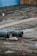 A construction site featuring large metal pipes resting on the ground, with red barriers scattered across the area. In the background, a blue tarp and wooden panels form a temporary wall. The ground is bare and appears recently disturbed.