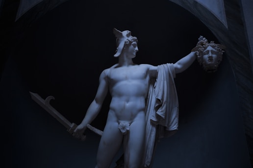 A white marble statue of a muscular figure wearing a winged helmet, holding a large sword in one hand and a severed head in the other. The statue is draped with a cloth over its left shoulder and the scene is set against a dark, arching background.