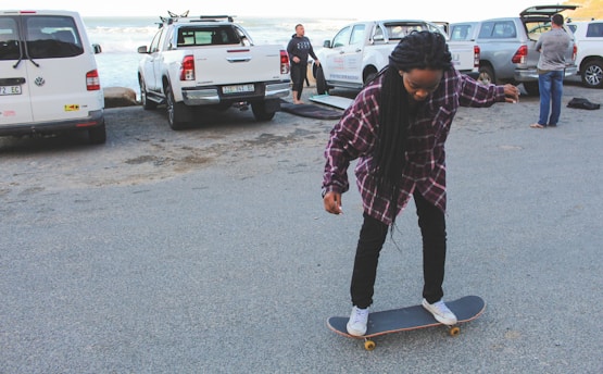 A person is skateboarding in a parking area near the beach. There are several parked cars, including pickup trucks and vans, and some people are standing near the vehicles. The person on the skateboard is wearing a plaid shirt and jeans.