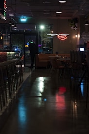 A dimly lit bar or restaurant interior with a reflective floor, empty chairs lined up along a counter, and a small seating area with tables. A neon sign depicting lips is on the wall, and a person stands near a glass door, appearing to look outside where colorful lights are visible.