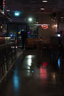 A dimly lit bar or restaurant interior with a reflective floor, empty chairs lined up along a counter, and a small seating area with tables. A neon sign depicting lips is on the wall, and a person stands near a glass door, appearing to look outside where colorful lights are visible.