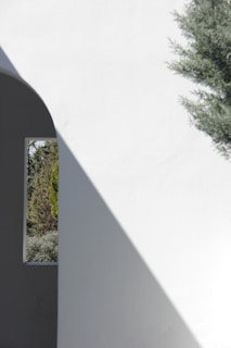 A minimalist architectural scene with a white wall casting a diagonal shadow, a small window revealing greenery outside, and a tree partially in view.