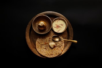 A wooden tray holds a traditional Indian meal. There is a paratha flatbread topped with butter, a small metallic bowl containing a brown dessert resembling a modak, and another bowl filled with a creamy white substance garnished with saffron strands. A brass spoon is placed on the paratha.