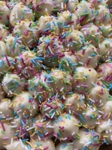 A close-up view of cookies or pastries topped with colorful sprinkles. The treats appear to be bite-sized and are coated with a white icing, adorned with an array of multicolored sprinkles.