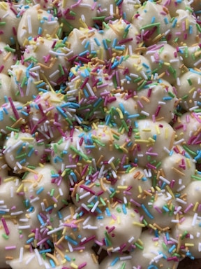 A close-up view of cookies or pastries topped with colorful sprinkles. The treats appear to be bite-sized and are coated with a white icing, adorned with an array of multicolored sprinkles.