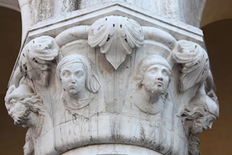 Carved stone pillar featuring intricate bas-relief sculptures of human faces and decorative foliage. The faces appear to be from varying historical or mythical figures, with detailed expressions and attire, suggesting an artistic or historical theme.