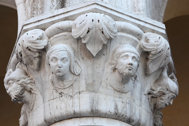 Carved stone pillar featuring intricate bas-relief sculptures of human faces and decorative foliage. The faces appear to be from varying historical or mythical figures, with detailed expressions and attire, suggesting an artistic or historical theme.