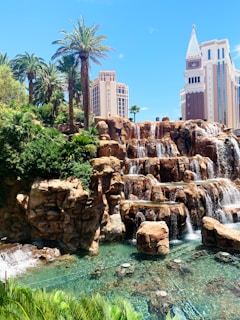 A picturesque scene featuring a cascading waterfall set against a background of lush greenery and palm trees. Two prominent buildings, labeled Palazzo and Venetian, rise majestically in the distance. The water below the waterfall is clear, with rock formations adding to the natural appeal.