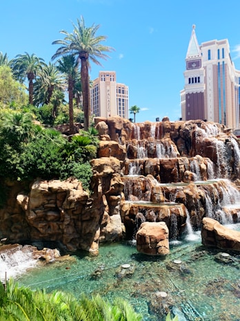 A picturesque scene featuring a cascading waterfall set against a background of lush greenery and palm trees. Two prominent buildings, labeled Palazzo and Venetian, rise majestically in the distance. The water below the waterfall is clear, with rock formations adding to the natural appeal.