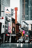 A busy urban street scene with various shops, including EB Games and Ed Mirvish Theatre. The street is wet with rain, and cars are driving past. Tall modern buildings surround older brick architecture, contributing to a mix of styles. Vibrant neon signs and billboards advertise theater shows and events, adding a lively atmosphere.