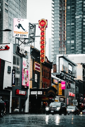 A busy urban street scene with various shops, including EB Games and Ed Mirvish Theatre. The street is wet with rain, and cars are driving past. Tall modern buildings surround older brick architecture, contributing to a mix of styles. Vibrant neon signs and billboards advertise theater shows and events, adding a lively atmosphere.