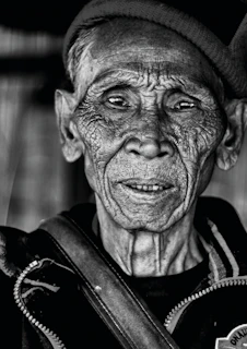 An elderly man with a weathered face looking out from a window, symbolizing resilience.