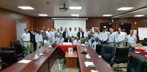 A group of people, mostly men, stand together in a conference or meeting room equipped with long tables and office chairs. They are gathered around a table with a large cake on it, suggesting a celebration or corporate event. The individuals are dressed in similar workwear, indicating they might be colleagues. The room has a formal setting, with a projector screen at the front.