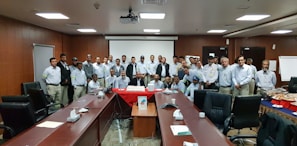 A group of people, mostly men, stand together in a conference or meeting room equipped with long tables and office chairs. They are gathered around a table with a large cake on it, suggesting a celebration or corporate event. The individuals are dressed in similar workwear, indicating they might be colleagues. The room has a formal setting, with a projector screen at the front.