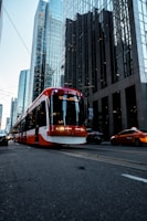 A red and white streetcar is traveling down a city street lined with tall glass skyscrapers. There is a taxi nearby and several other cars can be seen. The buildings reflect the light, adding a blue tint to the scene.