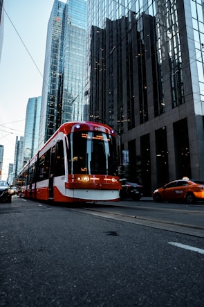 A red and white streetcar is traveling down a city street lined with tall glass skyscrapers. There is a taxi nearby and several other cars can be seen. The buildings reflect the light, adding a blue tint to the scene.
