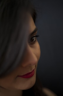 A close-up of a woman's face with focus on her eye and red lipstick. Her dark hair partially covers one eye, and the background is a soft blur, enhancing the intimate and mysterious atmosphere.