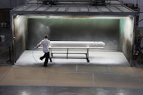A person is applying a spray finish to a long object in a contained spray booth. The environment appears industrial, with machinery and safety equipment visible. The lighting highlights the object being worked on, which is supported by a rolling metal frame.