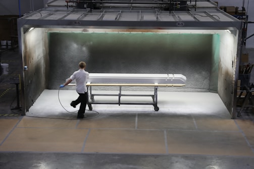 A person is applying a spray finish to a long object in a contained spray booth. The environment appears industrial, with machinery and safety equipment visible. The lighting highlights the object being worked on, which is supported by a rolling metal frame.