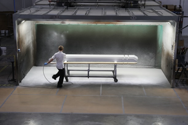 A person is applying a spray finish to a long object in a contained spray booth. The environment appears industrial, with machinery and safety equipment visible. The lighting highlights the object being worked on, which is supported by a rolling metal frame.