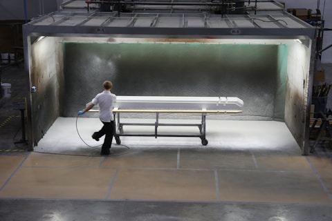 A person is applying a spray finish to a long object in a contained spray booth. The environment appears industrial, with machinery and safety equipment visible. The lighting highlights the object being worked on, which is supported by a rolling metal frame.