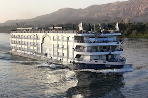 A large river cruise ship is navigating through a wide river with lush greenery on the banks and rugged, barren hills in the background. The vessel has multiple decks with ornate railings, and people can be seen enjoying the view from the top deck under umbrellas. The water is calm, reflecting the ship's movement.