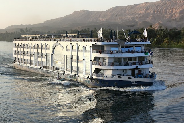 A large river cruise ship is navigating through a wide river with lush greenery on the banks and rugged, barren hills in the background. The vessel has multiple decks with ornate railings, and people can be seen enjoying the view from the top deck under umbrellas. The water is calm, reflecting the ship's movement.