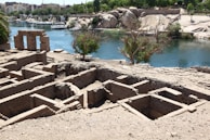 An archaeological site with ancient ruins consisting of rectangular brick foundations located near a calm blue river. In the background, lush green trees and modern buildings are visible, contrasting with the sandy, rocky terrain of the site. A large stone structure with three columns stands prominently by the water's edge.