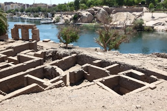 An archaeological site with ancient ruins consisting of rectangular brick foundations located near a calm blue river. In the background, lush green trees and modern buildings are visible, contrasting with the sandy, rocky terrain of the site. A large stone structure with three columns stands prominently by the water's edge.