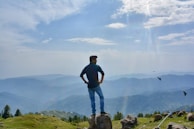 A person stands confidently on a rock, looking out over a vast mountainous landscape under a partly cloudy sky. Birds are seen flying nearby, and the scene conveys a sense of freedom and adventure.