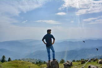 A person stands confidently on a rock, looking out over a vast mountainous landscape under a partly cloudy sky. Birds are seen flying nearby, and the scene conveys a sense of freedom and adventure.