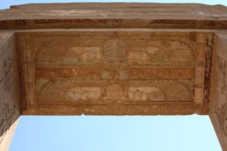 An ancient stone ceiling features intricate carvings, exhibiting detailed Egyptian hieroglyphs and symbolic motifs such as a winged sun disk. The stonework displays a blend of natural hues, creating a historical ambiance.