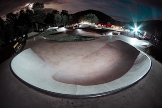A skateboard park under dramatic nighttime lighting, with smooth concrete curves and ramps forming a winding path. Trees line the background, illuminated by bright spotlights, creating a striking contrast with the cloudy sky. Benches and various equipment are visible along the sides.