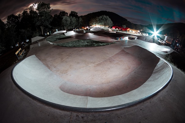 A skateboard park under dramatic nighttime lighting, with smooth concrete curves and ramps forming a winding path. Trees line the background, illuminated by bright spotlights, creating a striking contrast with the cloudy sky. Benches and various equipment are visible along the sides.