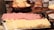 A variety of sliced meats and cheeses are neatly arranged on a platter. In the background, there are baskets of sliced bread and a loaf of packaged bread next to jars. Utensils and some scattered packets are nearby on the wooden table.