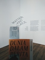 A gallery wall features a text display with the words 'Matter and Place' prominently displayed at an angle. Below, there is a person holding a book or booklet titled 'Dunia Dalam Berita' with an orange cover. The flooring is wooden and the overall atmosphere is minimalistic.