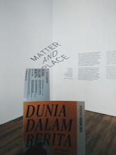A gallery wall features a text display with the words 'Matter and Place' prominently displayed at an angle. Below, there is a person holding a book or booklet titled 'Dunia Dalam Berita' with an orange cover. The flooring is wooden and the overall atmosphere is minimalistic.