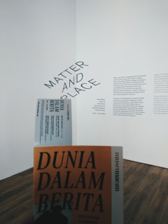 A gallery wall features a text display with the words 'Matter and Place' prominently displayed at an angle. Below, there is a person holding a book or booklet titled 'Dunia Dalam Berita' with an orange cover. The flooring is wooden and the overall atmosphere is minimalistic.