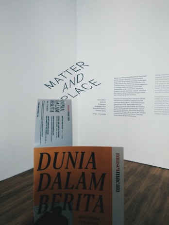 A gallery wall features a text display with the words 'Matter and Place' prominently displayed at an angle. Below, there is a person holding a book or booklet titled 'Dunia Dalam Berita' with an orange cover. The flooring is wooden and the overall atmosphere is minimalistic.