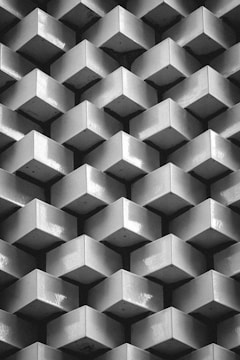 A repetitive pattern of three-dimensional, geometric blocks arranged in a grid. Each block appears as a cube protruding outward, creating an illusion of depth and texture. The image is in black and white, emphasizing the play of light and shadow across the surfaces.