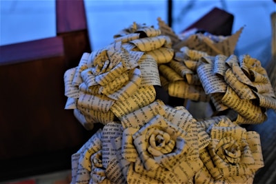 Intricately crafted paper roses made from printed text pages arranged in a bouquet. The background features a blurred, dark wooden structure and a soft blue hue. The roses have a delicate, artistic appearance with visible text adding texture.