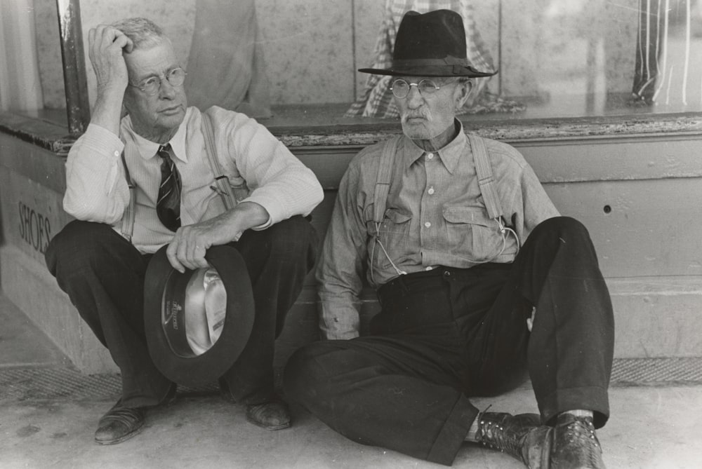 grayscale photo of two men sitting and leaning on wall