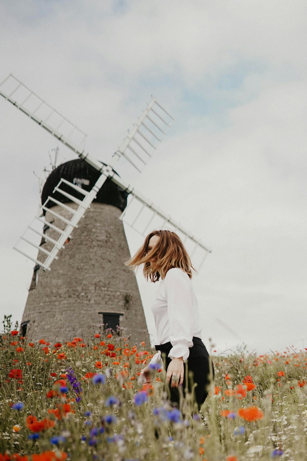 woman in white top and black pants standing in front of windmill at the flower field